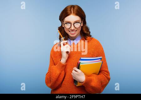 joyeux jeune étudiant en lunettes et chandail orange tenant des carnets et crayon isolé sur bleu Banque D'Images