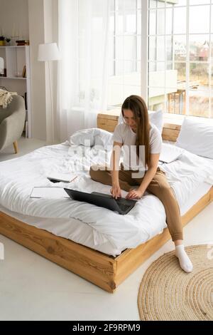 Femme en T-shirt blanc assis avec un ordinateur portable sur le lit à accueil Banque D'Images
