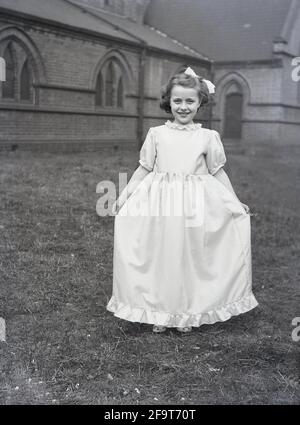 1956, historique, à l'extérieur dans le domaine d'une église, une jolie jeune fille montrant sa longue robe et robe qu'elle portera pour la traditionnelle procession de la Reine de mai, Angleterre, Royaume-Uni. May Day est un festival ancien pour célébrer l'arrivée du printemps, y compris le couronnement d'une reine de mai et la danse autour d'un Maypole, activités qui ont eu lieu en Angleterre pendant des siècles. Choisis parmi les filles de la région pour diriger le défilé du jour de mai, la reine de mai portant une couronne commencerait les célébrations et dans le nord de l'Angleterre, les écoles du dimanche de l'Église ont souvent dirigé l'organisation de la journée. Banque D'Images