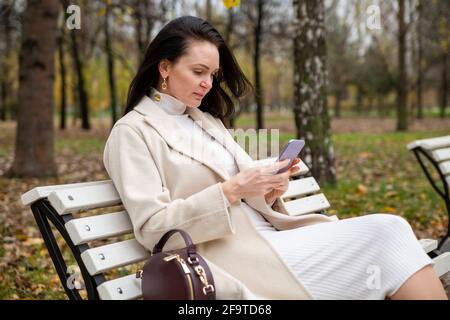 belle femme est en train de taper dans un smartphone et s'assoit sur un banc dans le parc. blogger crée du contenu. Internet surfing. Rencontres internet. Banque D'Images