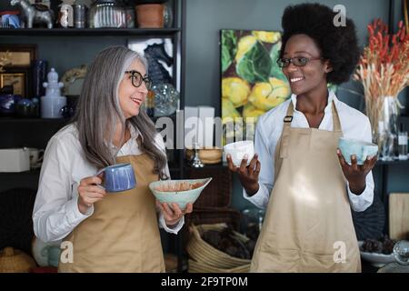 Souriantes femmes multiraciales dans des lunettes et un tablier tenant la décoration et regardant l'une l'autre. Deux femmes vendeurs travaillent ensemble dans un magasin moderne. Banque D'Images