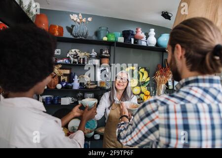 Vue arrière d'un jeune couple multiracial en vêtements décontractés achetant des articles à la boutique de décoration. Agréable femme de vente âgée dans les lunettes et le tablier de consultation sur les produits. Banque D'Images