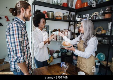 Jolie femme senior en tablier montrant aux jeunes clients des tasses et des assiettes en céramique exclusives au magasin de décoration. Vendeur compétent aidant le couple multiracial à choisir le produit. Banque D'Images