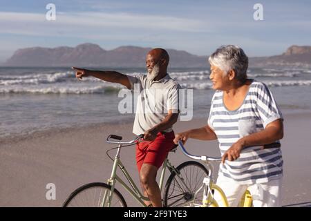 Couple afro-américain senior avec des vélos pointant vers une direction à la plage Banque D'Images