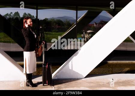 Belle jeune femme dans la rue dans la zone commerçante avec valise de voyage d'affaires parlant au téléphone. Concept d'appels d'affaires Banque D'Images