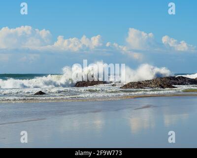 Journée de ciel bleu à la plage avec des vagues s'écrasant sur les rochers près du rivage devant des nuages blancs se reflétant sur le sable humide, Sawtell Australie Banque D'Images