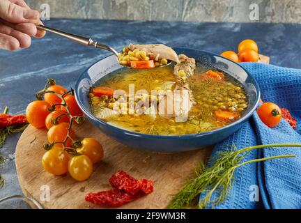 Soupe aux pois avec poulet, carottes, tomates et herbes dans une assiette profonde sur bois avec serviette sur fond bleu. Banque D'Images