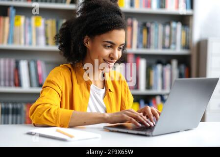 Une étudiante ou une freelance afro-américaine satisfaite, avec ses cheveux bouclés, habillée à la mode, est assise à un bureau, utilise un ordinateur portable, se concentre sur Internet, recherche d'informations, sourire Banque D'Images