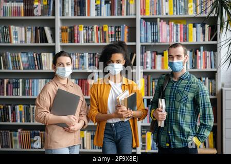 Éducation, vie étudiante, amitié. Des étudiants multinationaux amis, debout dans la bibliothèque de l'université, portant des masques médicaux protecteurs en raison de la pandémie du coronavirus, regardant la caméra Banque D'Images