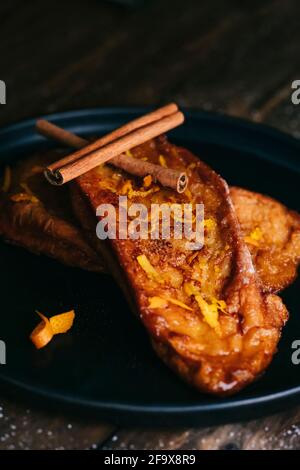 Vue rapprochée de deux tranches de pain frit avec deux bâtonnets de cannelle, de sucre et de citron et de pelures d'orange sur une assiette noire sur une table en bois sombre Banque D'Images