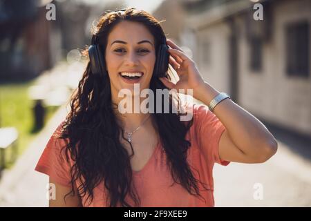 Une jeune femme heureuse profite d'une journée ensoleillée en plein air en ville. Elle écoute de la musique avec des écouteurs. Banque D'Images