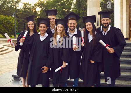 Étudiants diplômés en robes de chambre et en casquettes détenant des diplômes près du campus universitaire. Les jeunes célèbrent le commencement Banque D'Images