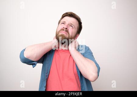 Homme caucasien en vêtements décontractés tient le cou avec les mains et regarde avec tristesse. Arrière-plan blanc. Banque D'Images