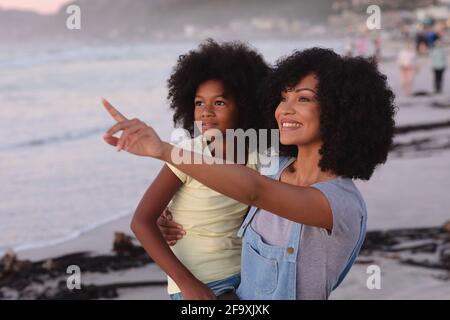 Souriante mère et fille afro-américaine embrassant à la plage, pointant vers l'avant Banque D'Images