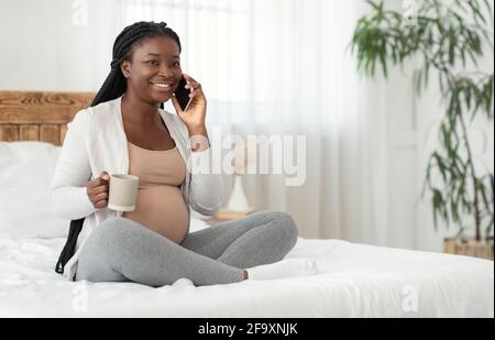 Passe-temps enceinte. Femme enceinte noire souriante parlant au téléphone portable et en train de boire du thé Banque D'Images