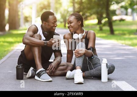 Un couple sportif noir souriant se repose avec un smartphone après un entraînement de fitness À l'extérieur Banque D'Images