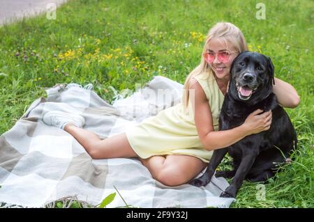 la jeune femme avec son grand chien noir est assise sur une couverture sur l'herbe dans une étreinte. Portrait de femme blonde en lunettes roses et chien Banque D'Images