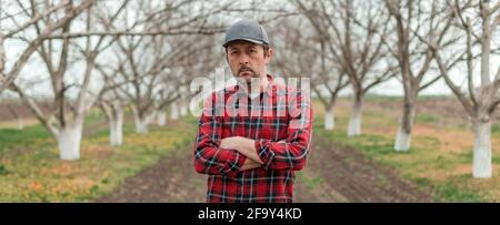 Portrait d'un beau fermier confiant avec casquette de baseball et chemise en flanelle debout avec les bras croisés dans le verger de fruits de noyer, foyer sélectif Banque D'Images