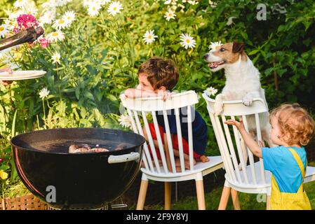 Les enfants et les chiens qui regardent la viande grillée à l'extérieur fête barbecue dans le jardin le jour d'été ensoleillé Banque D'Images