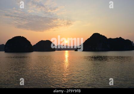 Vue panoramique du coucher de soleil sur les eaux étincelantes, les formations rocheuses, les montagnes bleues et les bateaux flottants sur la baie de Halong au Vietnam Banque D'Images