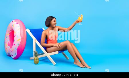 Bonne dame noire en maillot de bain tenant un cocktail, assise dans une chaise longue sur fond bleu, bannière avec espace libre Banque D'Images