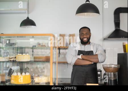 Souriant et sympathique propriétaire de petite entreprise afro-américaine, gérant de café, serveur de boulangerie multiracial se tient derrière le comptoir dans une posture confiante avec les bras croisés et regarde la caméra Banque D'Images