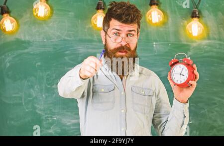 L'hipster barbu tient l'horloge, le tableau noir sur le fond. Homme avec la barbe et la moustache sur le visage surpris. Professeur de lunettes tient le réveil et Banque D'Images