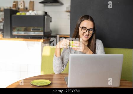 Une jeune femme souriante prend une pause de travail en ligne avec un ordinateur portable, assise à la réception dans un café confortable, tient une tasse avec café et regarde l'écran d'ordinateur Banque D'Images