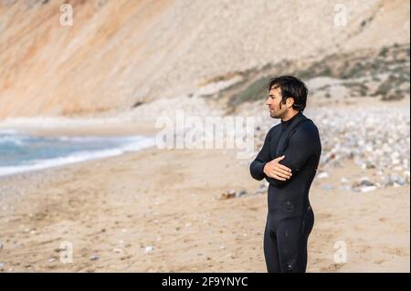 Vue latérale moyenne homme mouillé avec combinaison de plongée et bras pliés debout sur la plage et regardant la distance. Banque D'Images