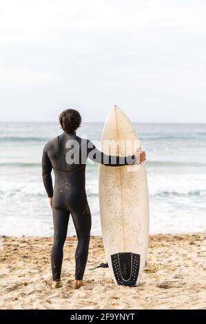 Vue arrière verticale de l'homme surfeur portant un costume de plongée incliné sur la planche de surf et debout en face de la mer regarder les vagues Banque D'Images