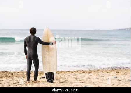 Vue arrière de l'homme surfeur portant un costume de plongée la planche de surf et se tenant en face de la mer regardant les vagues Banque D'Images