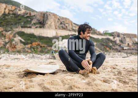 Jeune surfeur portant un costume de plongée assis sur le sable à côté de sa planche de surf souriant et se reposant après le surf. Banque D'Images