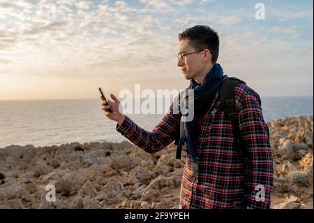 Jeune homme dans un endroit isolé sur une falaise rocheuse avec la mer sur fond. Homme tenant un smartphone et cherchant une connexion réseau. Banque D'Images