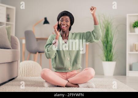 Photo pleine grandeur de la jeune femme africaine sourire heureux positif appel parler téléphone portable célébrer gagner victoire poing assis au sol intérieur Banque D'Images