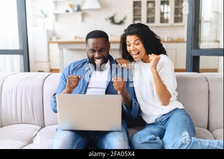 Un couple afro-américain hilarant célèbre le succès soulevé des poings serrés et criant heureusement, assis sur le canapé à la maison avec l'ordinateur portable sur les tours, gagné à la loterie, a obtenu la meilleure offre Banque D'Images