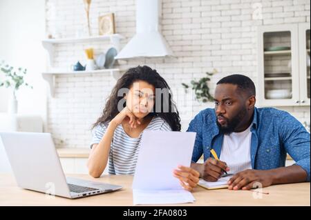 Couple multiracial calculant la finance, femme biracial et homme noir regardant à travers les papiers, assis à la table avec un ordinateur portable, prenant des notes, jeunes conjoints concentrés lisant le contrat hypothécaire Banque D'Images