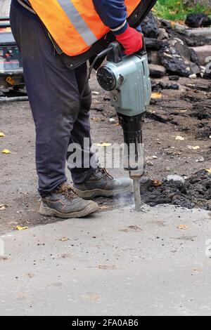 Un ouvrier de la route portant une veste réfléchissante éclate l'asphalte vieux de la route avec un marteau à inertie électrique. Image verticale, gros plan. Banque D'Images