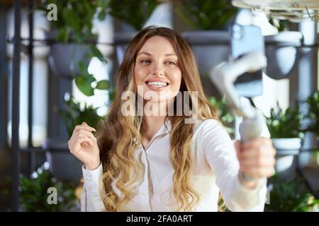 Portrait en face d'une jeune femme souriante et attrayante en chemise blanche tenant un bâton de selfie et communiquant avec les gens en direct. Concept de processus faisant de la vidéo ou selfie dans le café. Banque D'Images