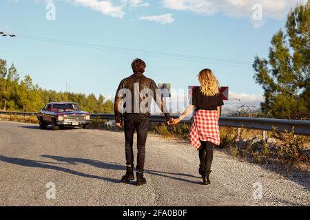 Vue arrière du couple portant des vêtements décontractés de style rock marchant vers leur muscle rouge vintage voiture garée sur le côté de la route sinueuse avec le bord Banque D'Images