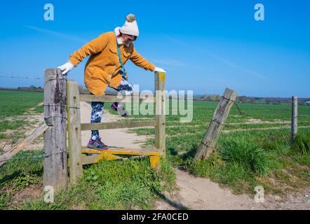 Femme âgée ou âgée vêtue d'un chapeau et d'un manteau marchant dans les champs et grimpant sur une caille en bois au printemps à West Sussex, Angleterre, Royaume-Uni. Banque D'Images