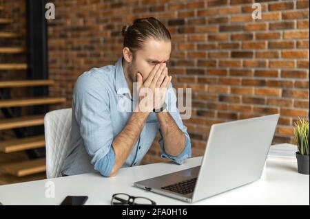 Un homme inquiet regarde l'écran d'un ordinateur portable, couvre le visage avec des palmiers, a trouvé une erreur dans un rapport, un employé de bureau ne parvient pas à terminer le travail avant la date limite Banque D'Images