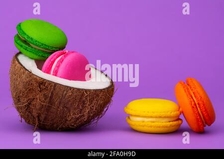 Macarons français posés dans des fruits de coco coupés en deux, sur fond violet clair. Banque D'Images