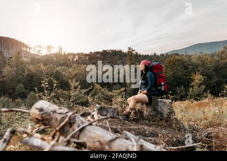 Une femme routard en admirant le coucher du soleil. Un portrait d'une femme qui fait une pause de la randonnée et de la détente sur une souche d'arbre. Banque D'Images
