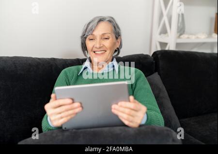 Une femme âgée et détendue, dans un chandail vert, se détend dans le salon, confortablement assise sur le canapé ou le canapé, regardant un spectacle de comédie sur une tablette ou un bloc-notes, lisant des nouvelles, parlant avec un ami Banque D'Images
