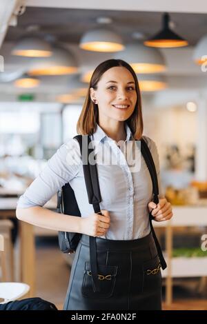 Photo d'intérieur de jeune européenne caucasienne isolée vêtue d'une chemise bleue avec sac à dos souriant happilyin un magasin, un restaurant, un bureau Banque D'Images