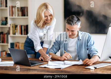 Discussion d'un projet d'entreprise. Deux partenaires d'affaires caucasiens d'âge moyen, heureux et agréables, vêtés de vêtements décontractés à la table, échangeant des idées, regardant attentivement les documents, souriant Banque D'Images