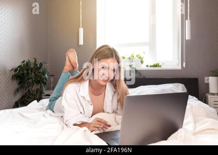 Jolie fille caucasienne souriante pose sur le lit dans la chambre et utilise un ordinateur portable pour le travail à distance. Communication, technologie, concepts de travail à distance dans un intérieur gris Banque D'Images