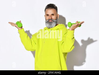 Portrait de l'homme barbu, vieux et brutal, en jaune sweat à capuche tenant les haltères vertes dans les mains sur les paumes ouvertes Banque D'Images