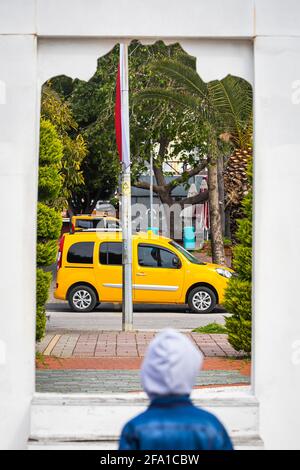 Concept de voyage. Un petit garçon regarde à travers une porte blanche, une fenêtre à un taxi jaune se déplaçant de l'autre côté de la rue vers des arbres verts, des palmiers dans un ho chaud Banque D'Images