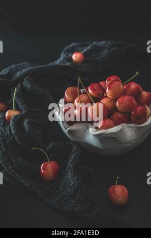 Cerises douces orange mûres de type « Anne royale » dans un bol blanc sur tissu noir et fond noir Banque D'Images